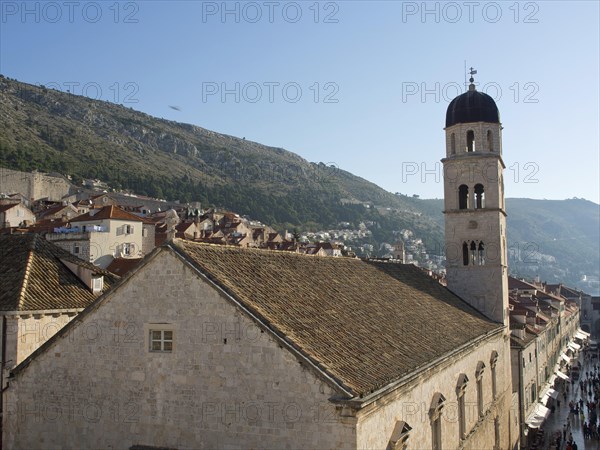 A bell tower rises above a church in a medieval town that stretches along a sunlit hill, the old town of Dubrovnik with historic houses, churches, red roofs and fortress walls, dubrovnik, coratia