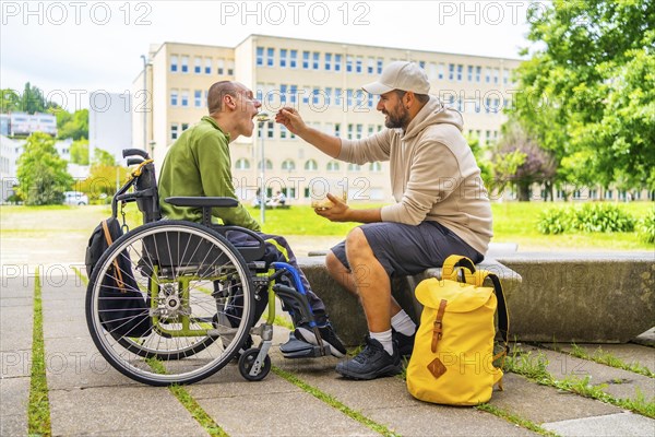 Full length caucasian male friend feeding a man with disability outside the university campus