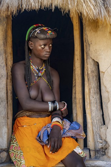 Hakaona woman with traditional kapapo hairstyle, sitting in the entrance of her mud hut, in the morning light, Angolan tribe of the Hakaona, near Opuwo, Kunene, Namibia, Africa