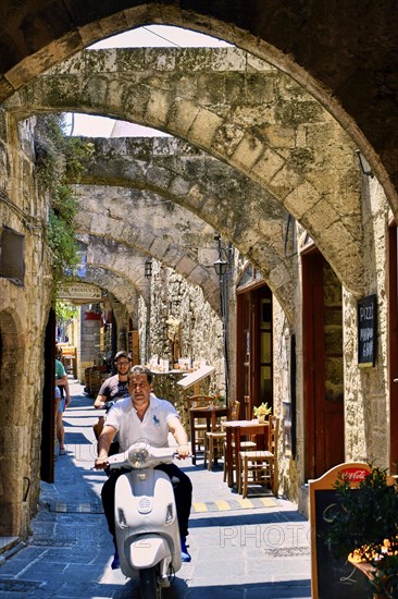Man riding a scooter through an old, sunny street with stone arches and historic buildings, Old Town of Rhodes, Rhodes Town, Rhodes, Dodecanese, Greek Islands, Greece, Europe