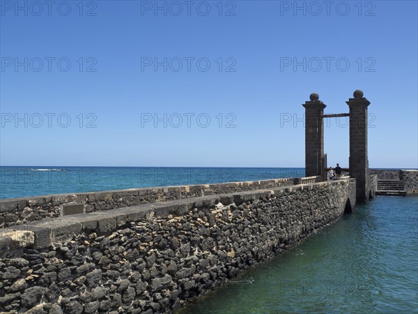A stone footbridge leads to the sea with a Tor tor in the foreground and blue water all around, arrecife, Lanzarote, Canary Islands, Spain, Europe