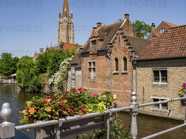 A canal with decorated balustrades and flowers along the row of brick-built houses in front of a church tower in sunny weather, Bruges, Belgium, Europe