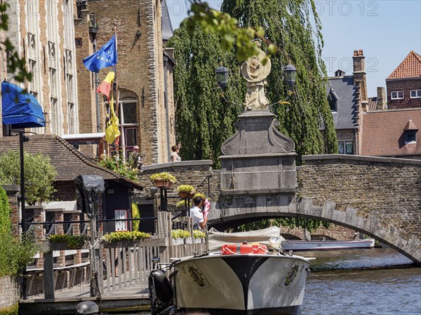 A boat under a bridge near historic buildings, decorated with flags and flowers, Bruges, Belgium, Europe