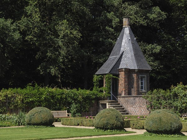 A small tower with a slate-covered roof in the middle of a well-tended garden with shrubs, green grass and trees providing shade, ochtrup, münsterland, germany