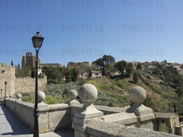 Picture of a bridge with a church in the background, surrounded by a green hilly landscape in bright sunlight, Toledo, Spain, Europe