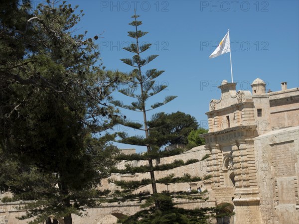 Historic fortress with stone wall, waving flag and tall trees against a blue sky, mdina, mediterranean sea, Malta, Europe