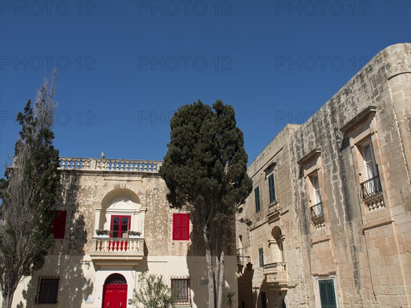 View of a square with historic buildings and red shutters, accompanied by trees under a cloudless sky, mdina, mediterranean sea, Malta, Europe