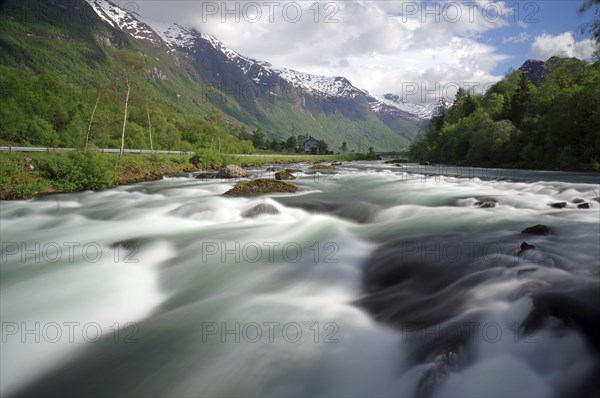 A wide river in a mountainous landscape with snow-capped peaks and cloudy skies, Olden, Nordfjord, Norway, Europe