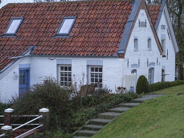 A white farmhouse with a red tiled roof and a staircase surrounded by grass and shrubs in a rural setting, greetsiel, east frisia, germany