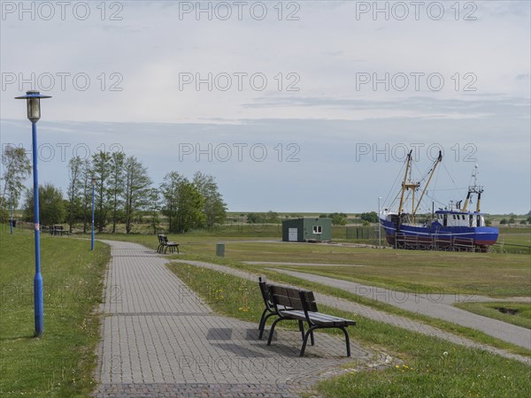 A park path with benches leads along a rural setting with fishing boats in the background, greetsiel, east friesland, germany