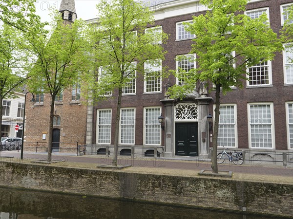 An old, historic brick building with many windows and trees, next to a quiet canal, Delft, Holland, Netherlands