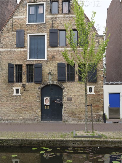 A historic brick building with black shutters, green trees and a quiet canal in the foreground, Delft, Holland, Netherlands