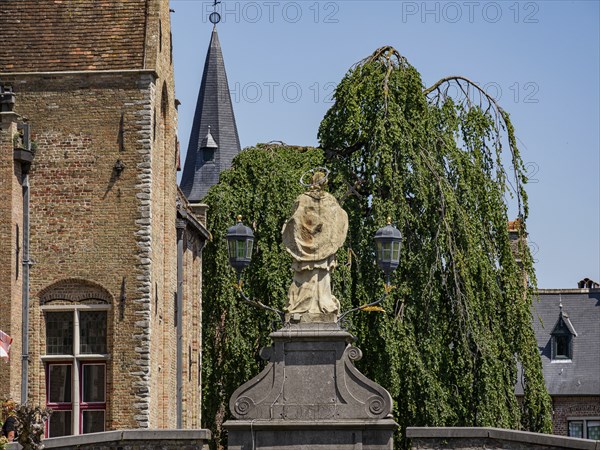 Statue in the foreground in front of historic brick buildings, a church with spire and gardens, Bruges, Flanders, Belgium, Europe