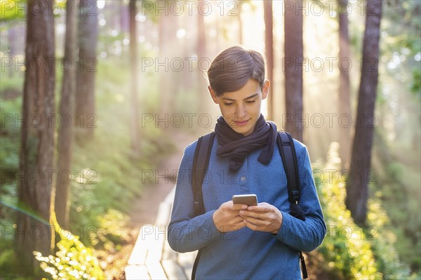 A boy stands on a forest path in the sunlight, carrying a rucksack and using his smartphone, AI generated, AI generated