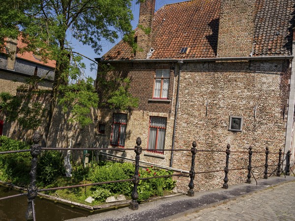 A historic building with a tiled roof and lush green trees along a canal in bright sunshine, Bruges, Belgium, Europe