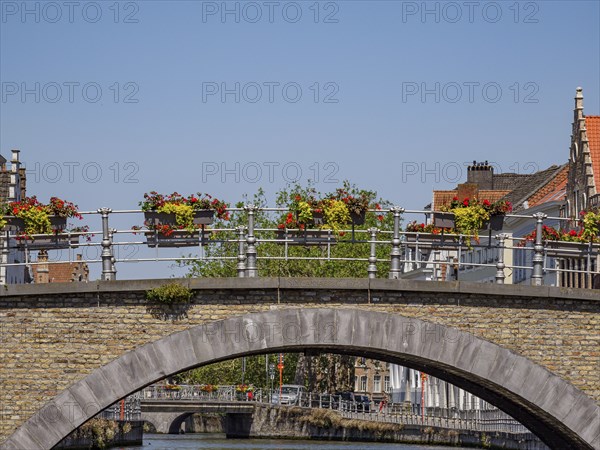A bridge decorated with flowers over a waterway, surrounded by historic buildings on a clear spring day, Bruges, Belgium, Europe