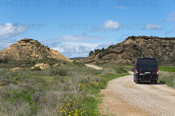 Black van on a gravel road in a hilly, green landscape under a blue sky with clouds, motorhome, camper, Bardenas Reales Natural Park, desert, semi-desert, Navarra, Nafarroa, UNESCO Biosphere Reserve, Spain, Europe
