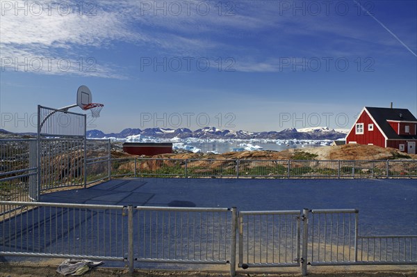 A basketball court on a coastline with a red house in the background and icebergs in the ocean, Tiniteqilaaq, East Greenland, Denmark, Europe