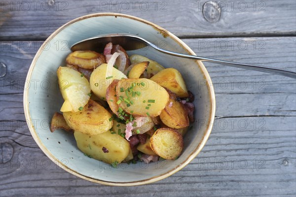 Fried potatoes with onions, bacon and chive garnish in a ceramic bowl on a blue gray wooden table, high angle view from above, copy space, selected focus, narrow depth of field