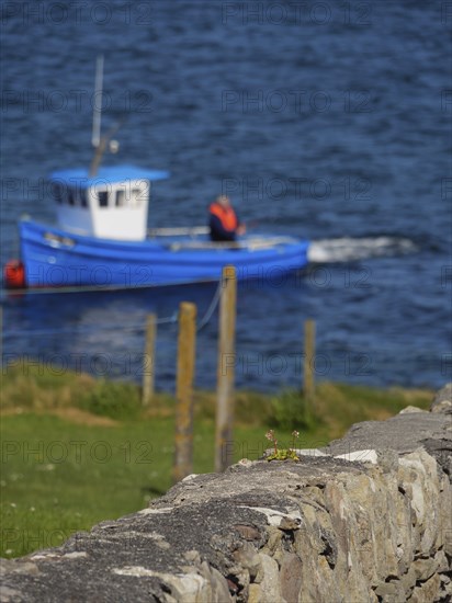 A blurred fishing boat and fence are seen behind a stone wall over blue water, Lerwick, Shetlands, Scotland, United Kingdom, Europe