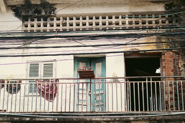 Facade of an old, dilapidated building with peeling paint, a balcony with hanging clothes, and numerous wires crisscrossing in front in the town of Kampot, Cambodia, Asia