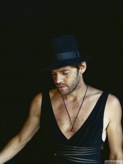 Man in a black hat and attire, wearing a necklace with red charm, appearing deep in thought under dim lighting, AI generated