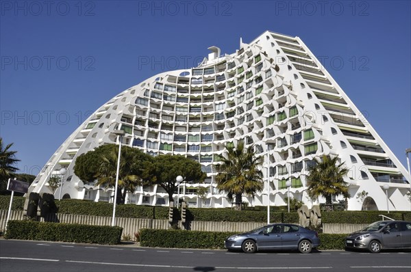 A modern curved building with a white facade and numerous balconies, set in an urban environment with trees and parked cars under a clear sky, Buildings in La Grande Motte