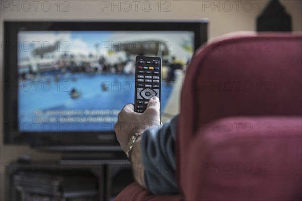 Man in comfy chair switching channels with remote control to look for pleasing programme on television set in living room