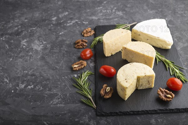Cheddar cheese and various types of cheese with rosemary and tomatoes on black slate board on a black concrete background. Side view, copy space, selective focus