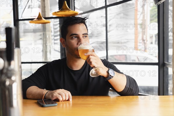 A young Latin man in a black shirt tastes a lager beer at a wooden bar table in a modern bar with stylish decor. The setting features unique lighting and a contemporary design, perfect for relaxation and socializing