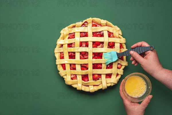 Woman hands washing with egg yolk the pie crust. Top view with strawberries and rhubarb tart. Uncooked berries pie
