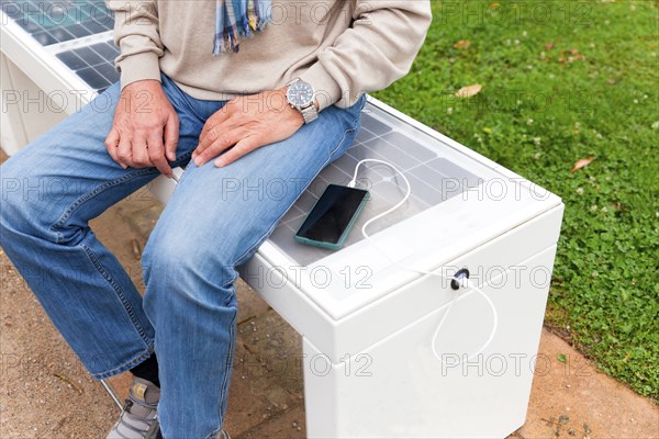 Man sits on a Smart Bench and charges his mobile phone, modern technology, bench with solar panel and mobile phone charger