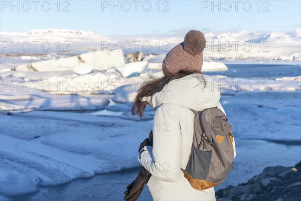 A woman wearing a white coat and a pink hat stands on a snowy beach looking out at the ocean