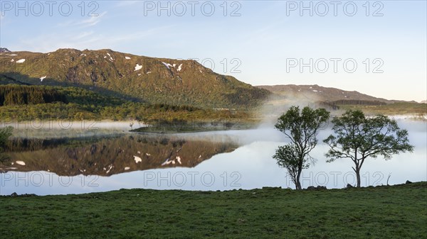 Landscape in the Lofoten Islands. Lake Lilandsvatnet, mountains in the background. Trees in the foreground. A light morning mist forms over the water. At night at the time of the midnight sun in good weather, blue sky. Golden hour. Reflection. Early summer. Vestvagoya, Lofoten, Norway, Europe