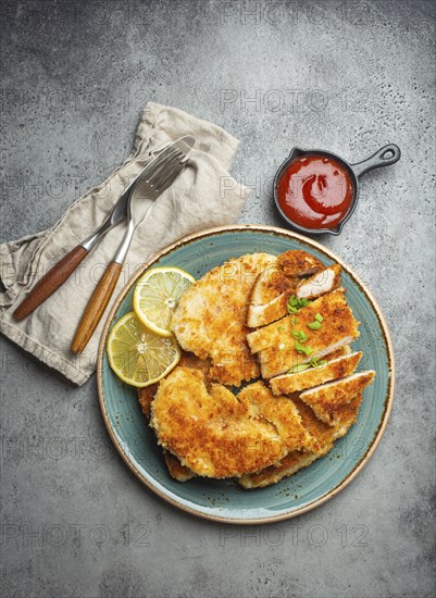 Crispy panko breaded fried chicken fillet with green salad and lemon cut on plate on gray rustic concrete background table with ketchup from above. Japanese style deep fried coated chicken breasts, food photography
