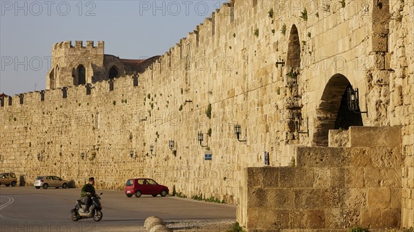Old city wall next to a road with passing scooter in sunshine, harbour area, Rhodes Town, Rhodes, Dodecanese, Greek Islands, Greece, Europe