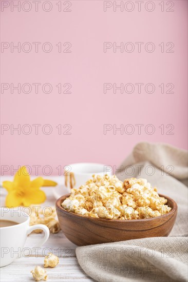 Popcorn with caramel in wooden bowl and a cup of coffee on a white and pink background and linen textile. Side view, copy space, selective focus