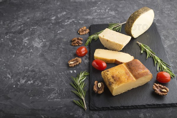 Smoked cheese and various types of cheese with rosemary and tomatoes on black slate board on a black concrete background. Side view, copy space, selective focus