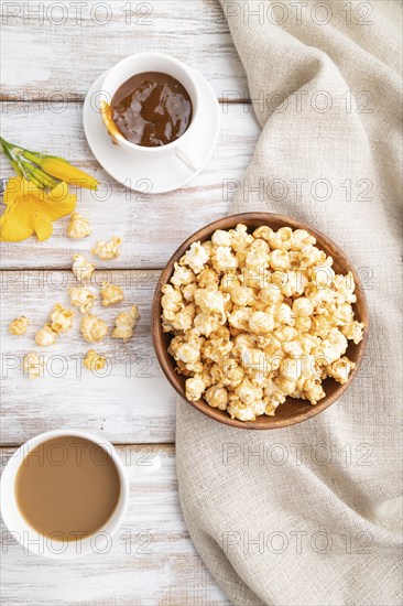 Popcorn with caramel in wooden bowl and a cup of coffee on a white wooden background and linen textile. Top view, flat lay, close up