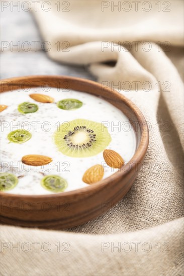 Yogurt with kiwi, gooseberry, chia and almonds in wooden bowl on gray wooden background and linen textile. Side view, close up, selective focus