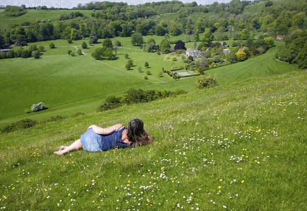 Woman lying on chalk grassland looking into deep green valley containing Rainscombe House, Oare Hill, Wiltshire, England, United Kingdom, Europe