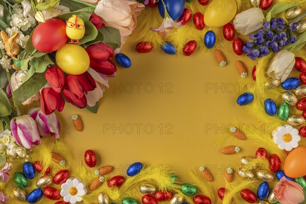 Colourful Easter frame with spring flowers, feathers, chocolate eggs and Easter eggs on a yellow background