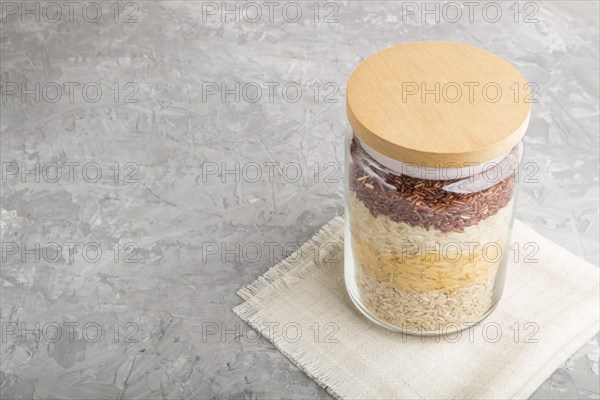Glass jar with different kinds of rice poured in layers on a gray concrete background. side view, close up, copy space. Healthy food concept