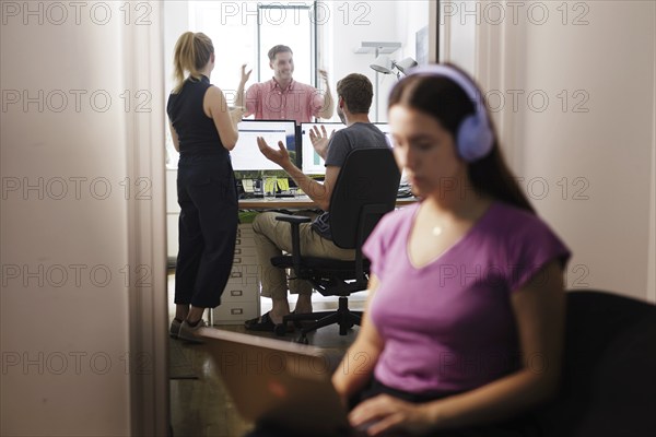 A woman works with headphones on her laptop while colleagues talk loudly. Berlin, 08.08.2024