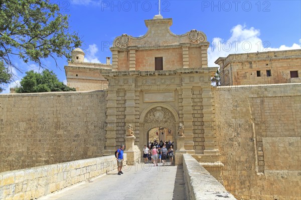 Tourists walking over moat bridge through entrance gateway of medieval city of Mdina, Malta, Europe