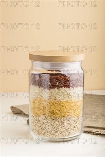 Glass jar with different kinds of rice poured in layers on white and orange background. side view, close up. Healthy food concept