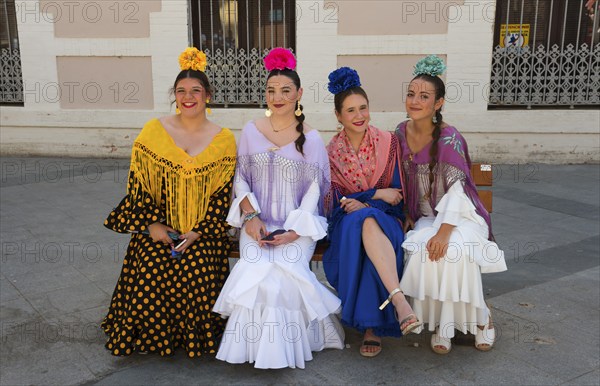 Four woman in colourful flamenco dresses and flowers in their hair sit on a bench on a street in traditional Spanish fashion, Feria de la Manzanilla, Feria de Sanlúcar, Sanlúcar de Barrameda, Sanlucar, Cádiz, Cadiz, Andalusia, Spain, Europe