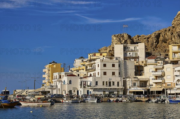 Whitewashed buildings in a coastal town at the harbour with boats and the Greek flag on a hill in the background, Pigadia, town and harbour, Pigadia Bay, main town, Karpathos, Dodecanese, Greek Islands, Greece, Europe