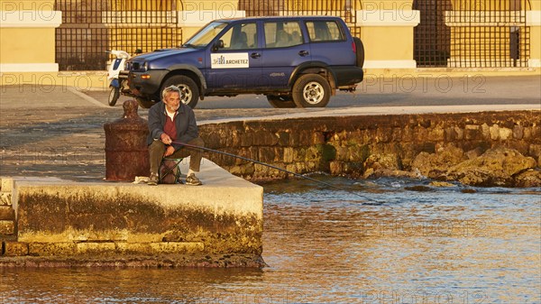 A man relaxes while fishing by the sea next to a parked off-road vehicle, Mandraki Oat, harbour area, Rhodes Town, Rhodes, Dodecanese, Greek Islands, Greece, Europe