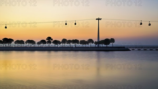 Silhouettes of trees and cable cars against a serene sunset reflecting on the calm water, Lisbon